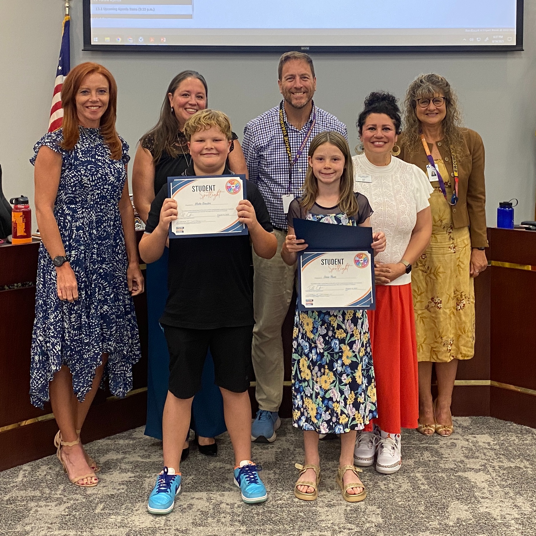 A male and a female student hold up their Student Spotlight certificates. They are surrounded by Board of Education members in the LPS Boardroom.