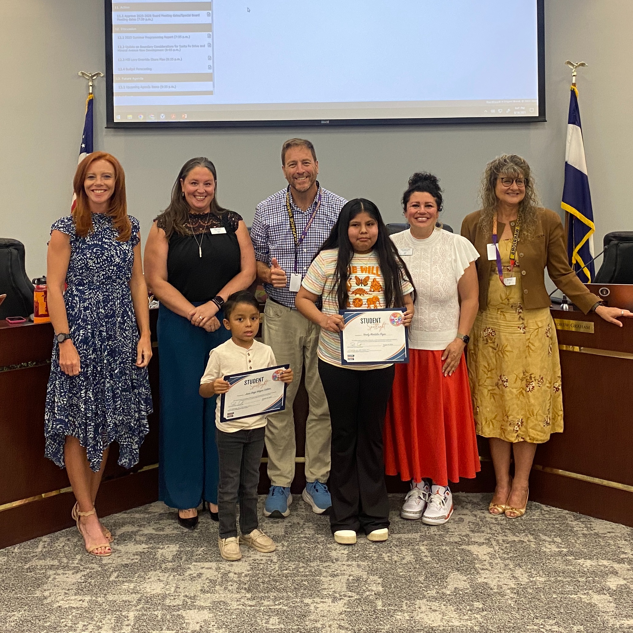 One male and one female student hold their Student Spotlight certificates. They are surrounded by the Board of Education in the LPS Boardroom.