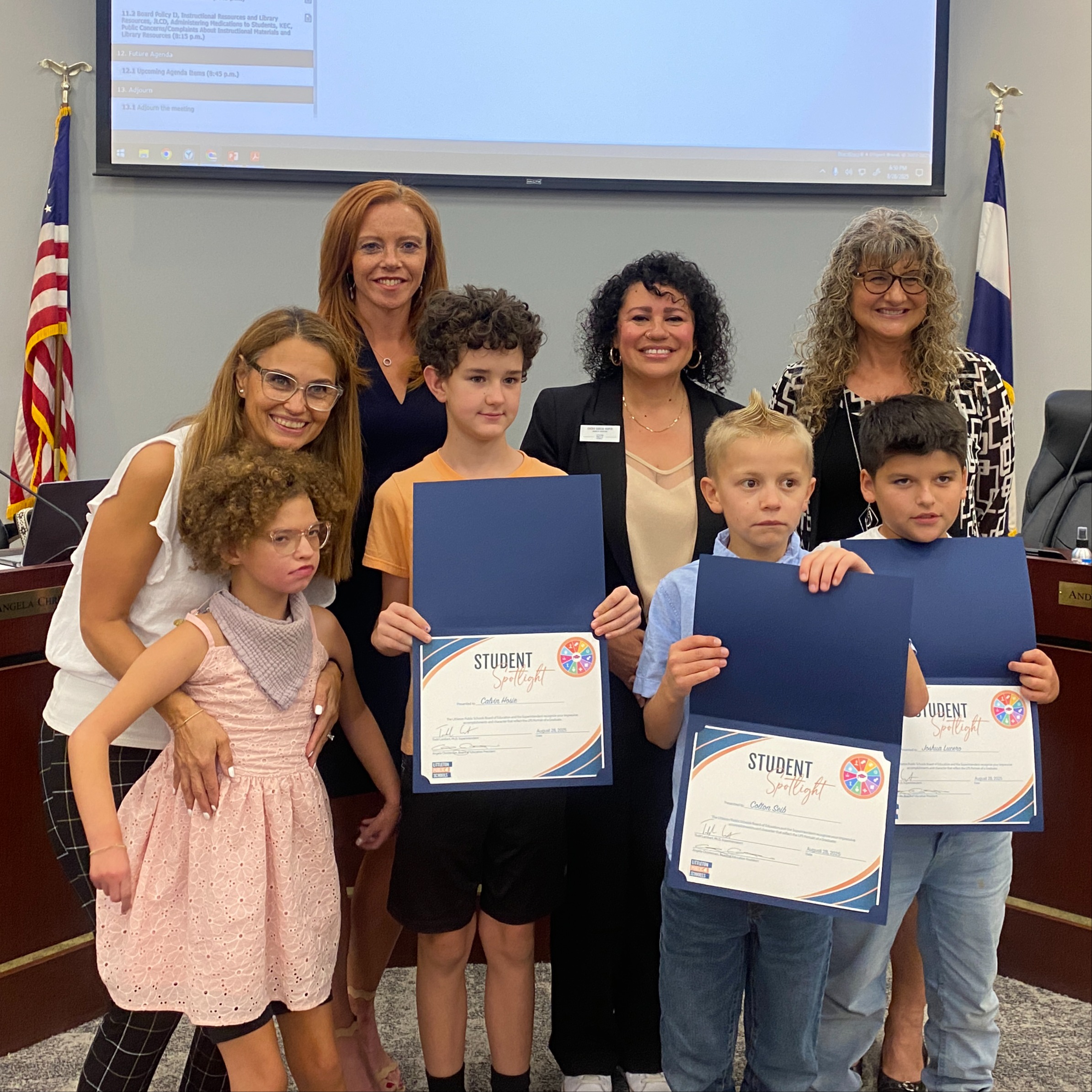 Four elementary students, three holding “Student Spotlight” certificates, pose with four smiling adults in a boardroom with flags and a screen.