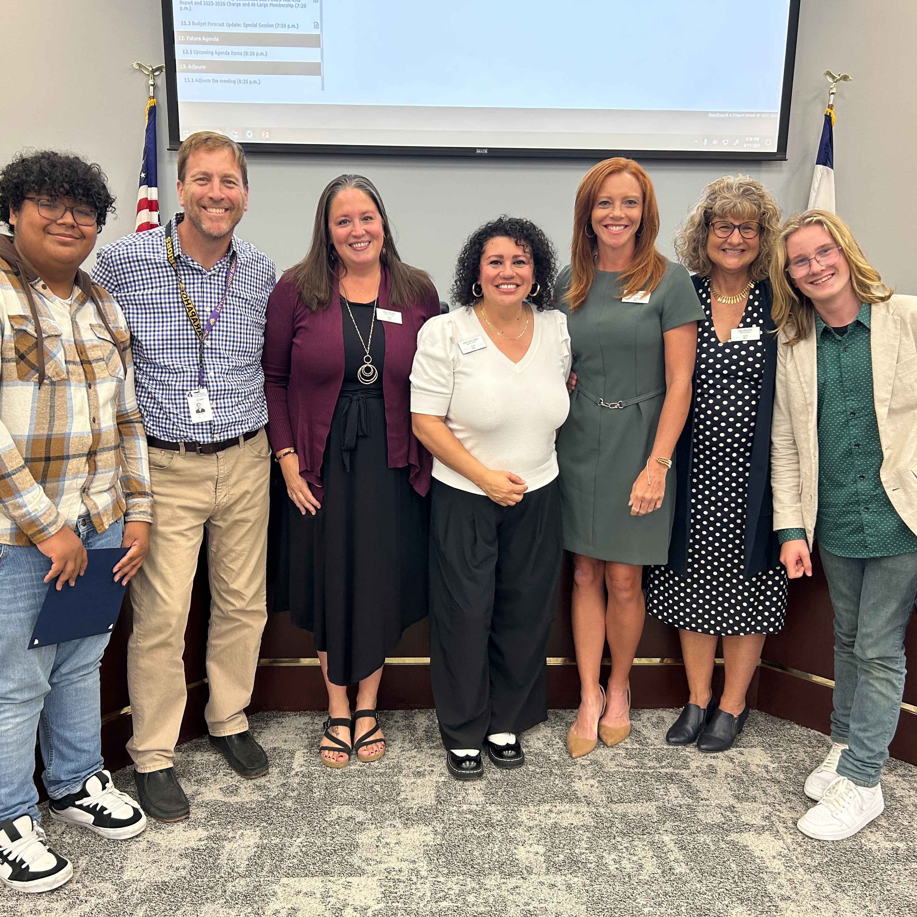 Two students stand at each end of a group photo with the Board of Education; diverse in age, gender, and ethnicity; flags and screen in background.