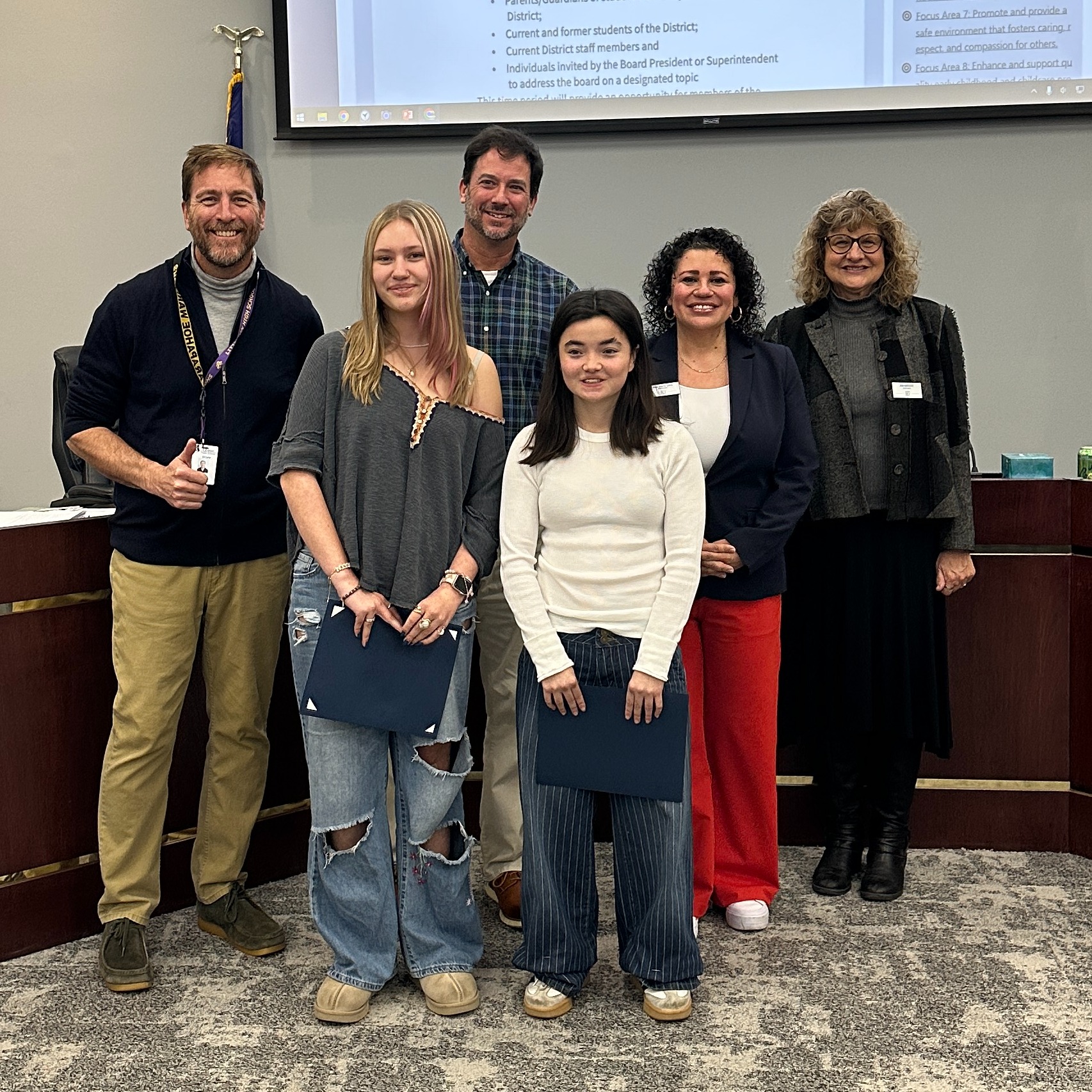Two students hold certificates while standing with four adults in front of a presentation screen at a board meeting.