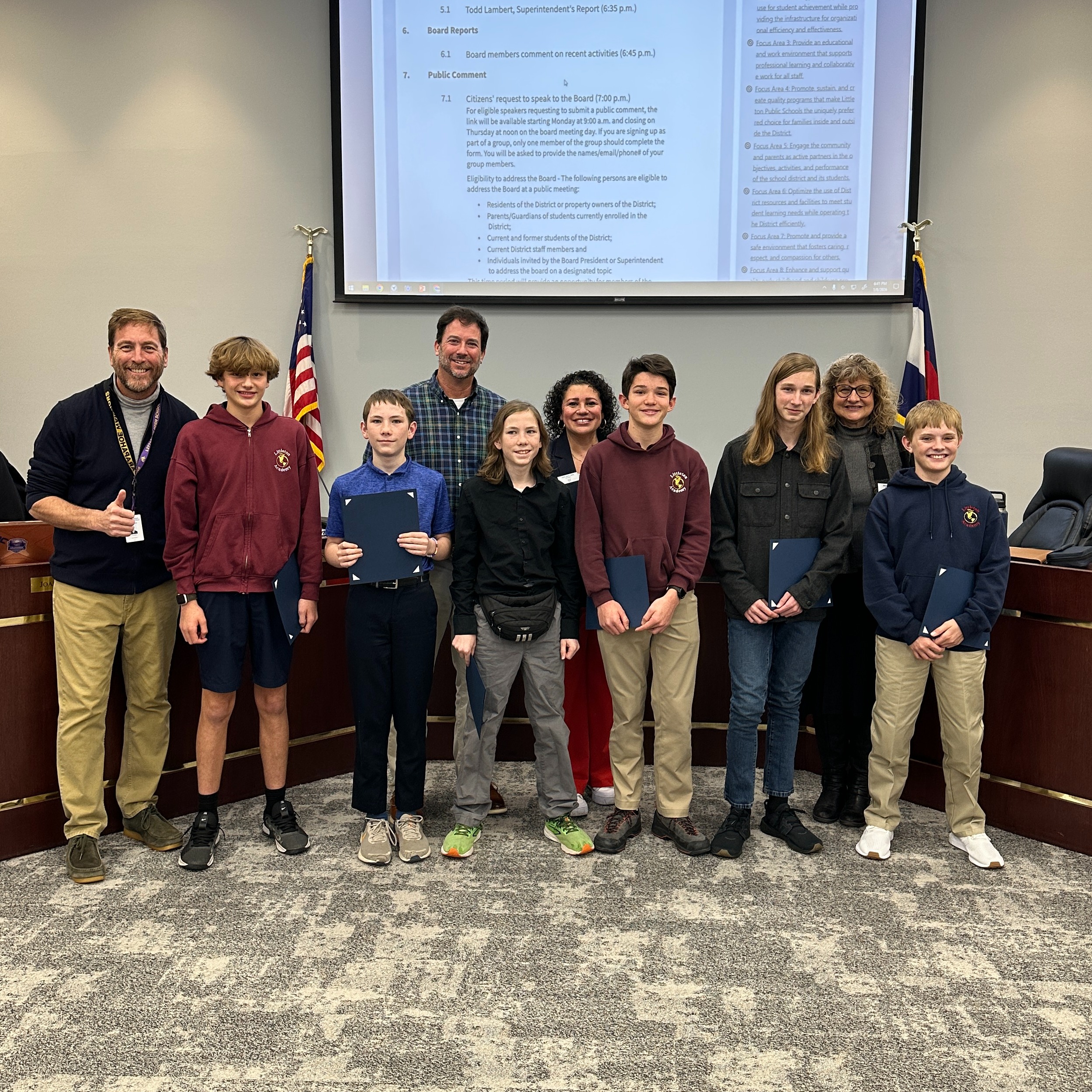 Six students hold awards and stand with four adults in front of a large screen at a Littleton Public Schools board meeting.