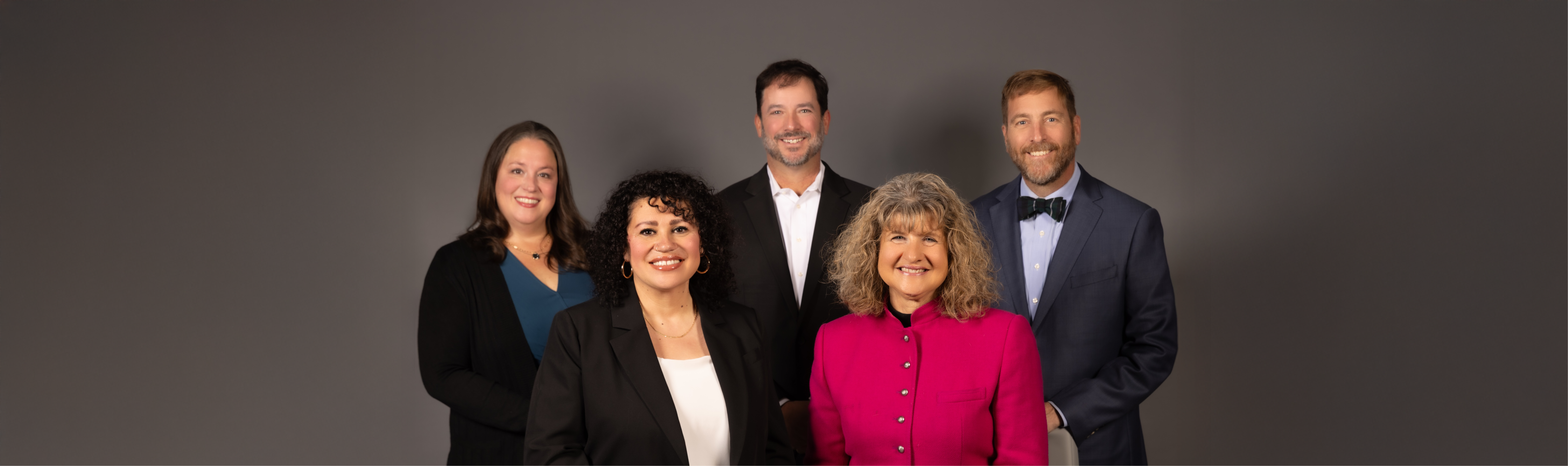 Group portrait of five Board of Education members posed in front of a textured backdrop. Three women stand in the back row and two individuals, one woman and one man, are seated in front. All are dressed professionally and smiling at the camera, representing district leadership in a formal and approachable manner.