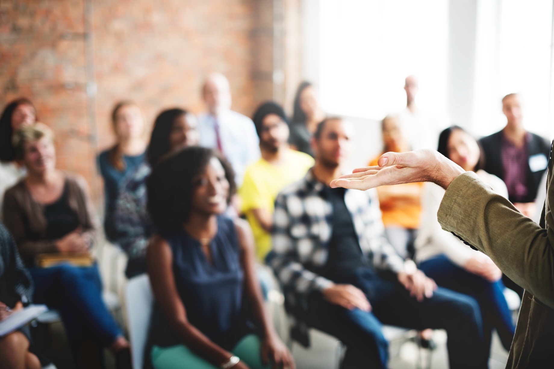 An audience of diverse adults sitting and listening to a presenter gesturing during a meeting or workshop.