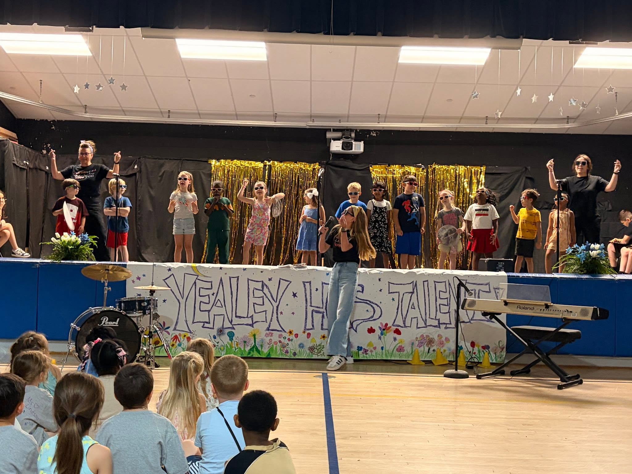 Stage with a banner. A person stands in front of a group on the stage. Audience seated in front.