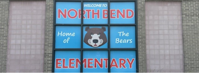 A sign outside of the school building. It has a blue background with red and white text that reads "Welcome to North Bend Elementary. Home of the Bears." In the center of the sign is a smiling bear face.
