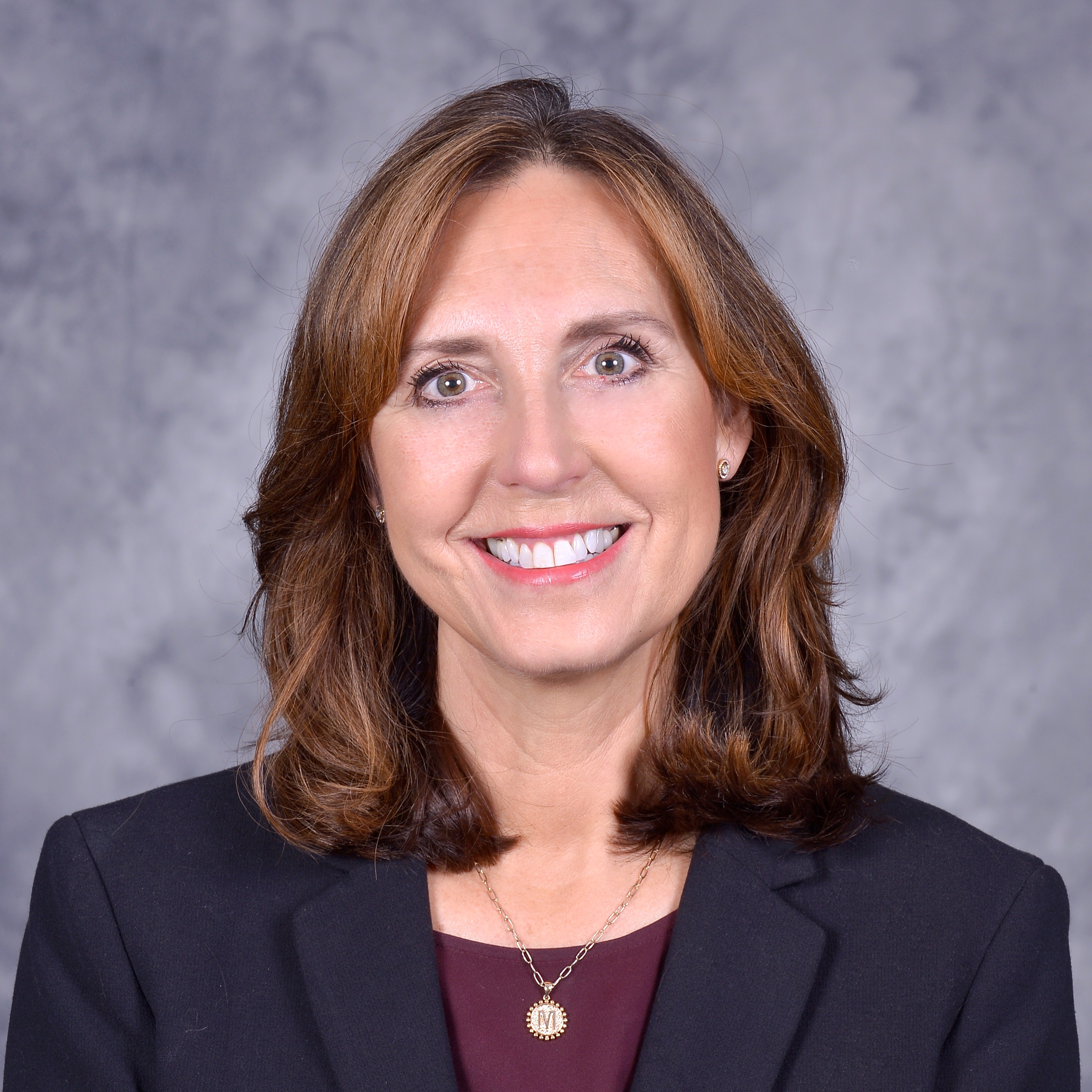 A professional headshot of a woman with shoulder-length brown hair, wearing a black blazer and smiling against a gray background.