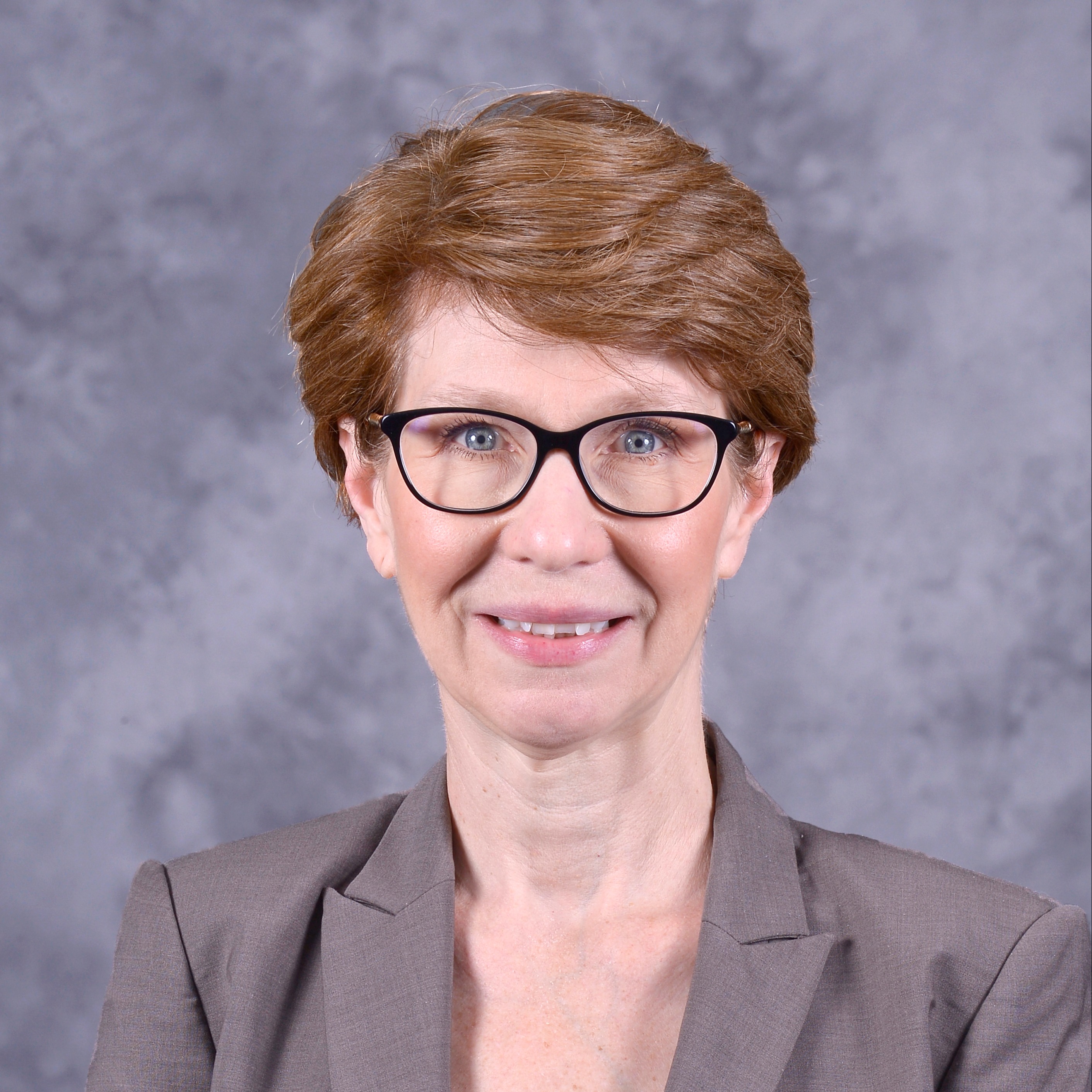 A woman with short brown hair and glasses smiles at the camera, wearing a black top against a gray background.