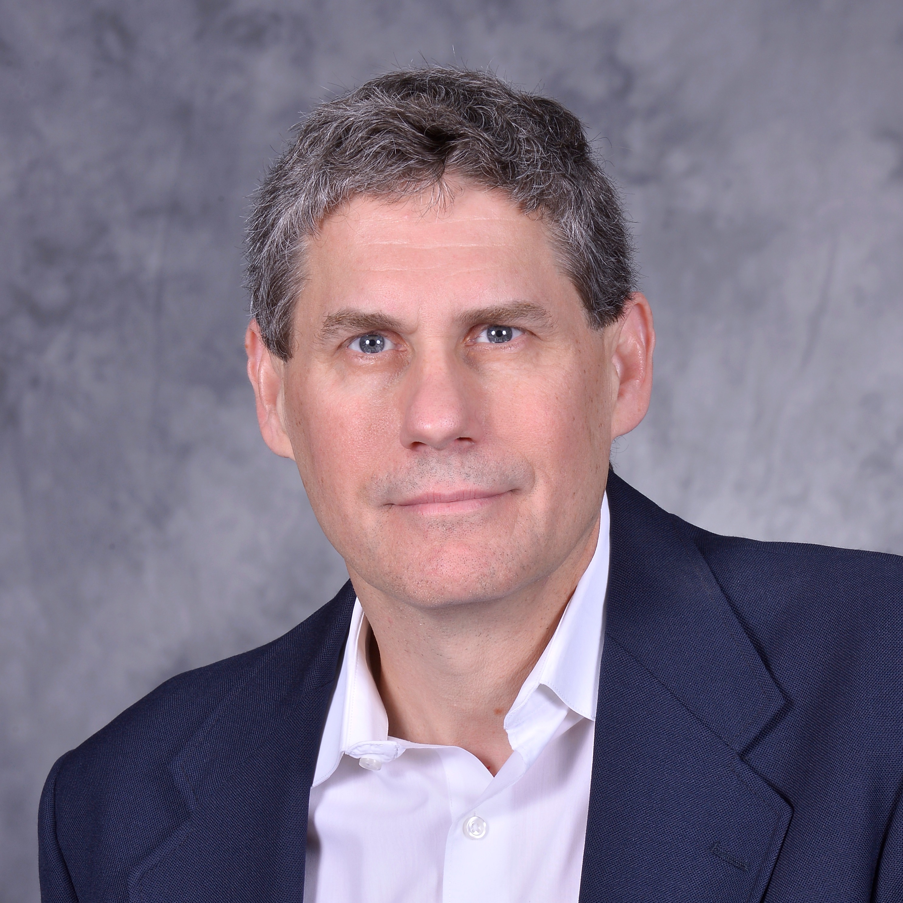 A man with short, curly hair wearing a black blazer over a white shirt, standing against a textured gray background.