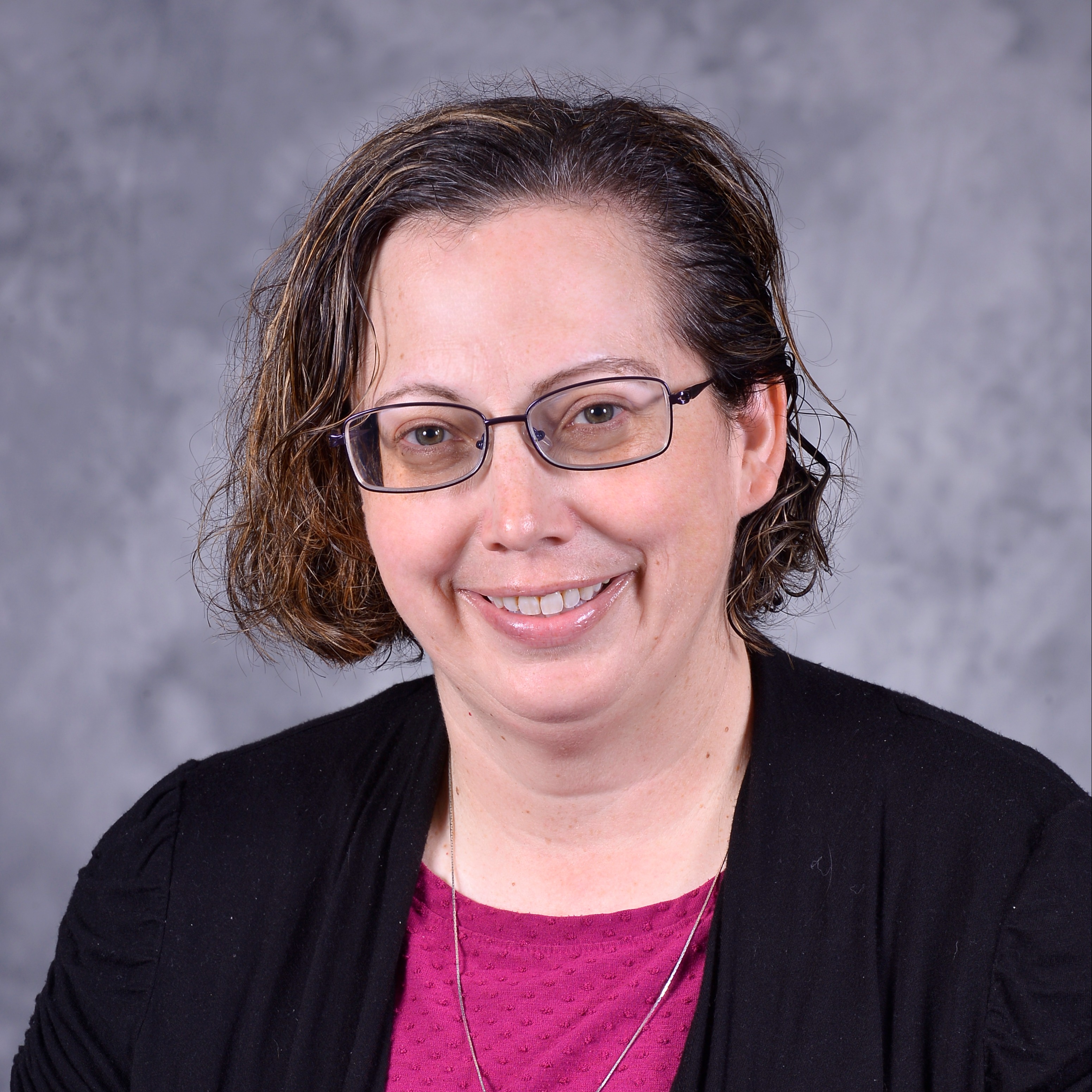 A woman with curly hair and glasses smiles while wearing a purple blouse and a cross necklace, set against a gray background.