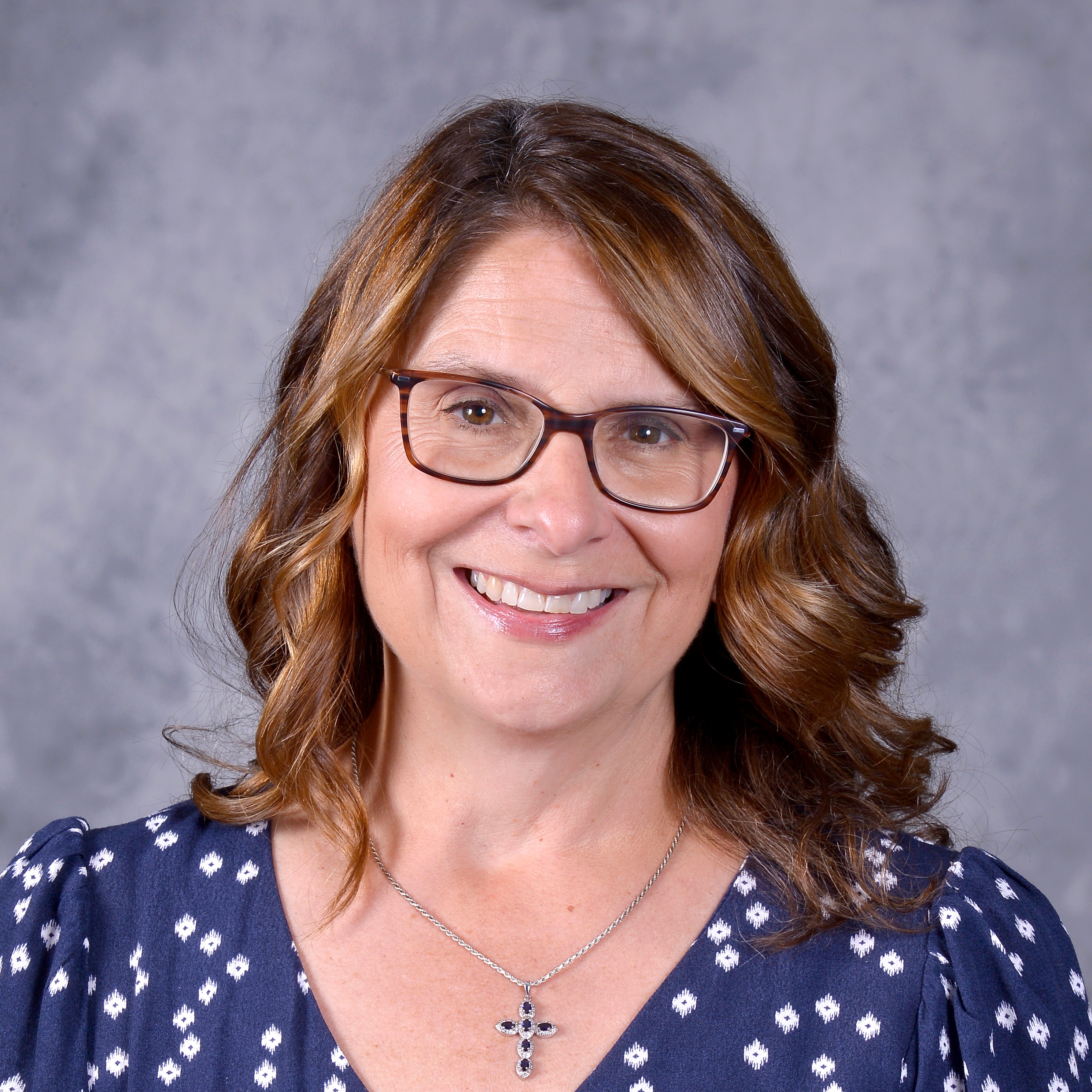 A woman with shoulder-length brown hair, wearing glasses and a floral dress, smiles warmly at the camera against a gray background.
