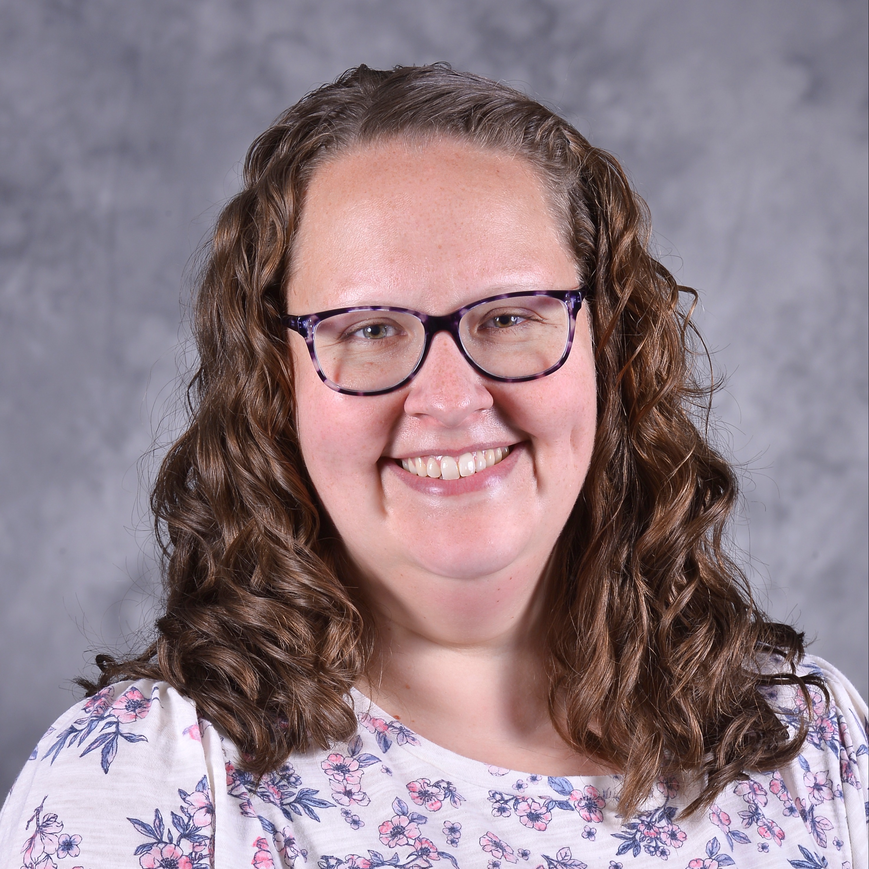 A smiling woman with glasses wearing a floral-patterned top, set against a textured gray background.