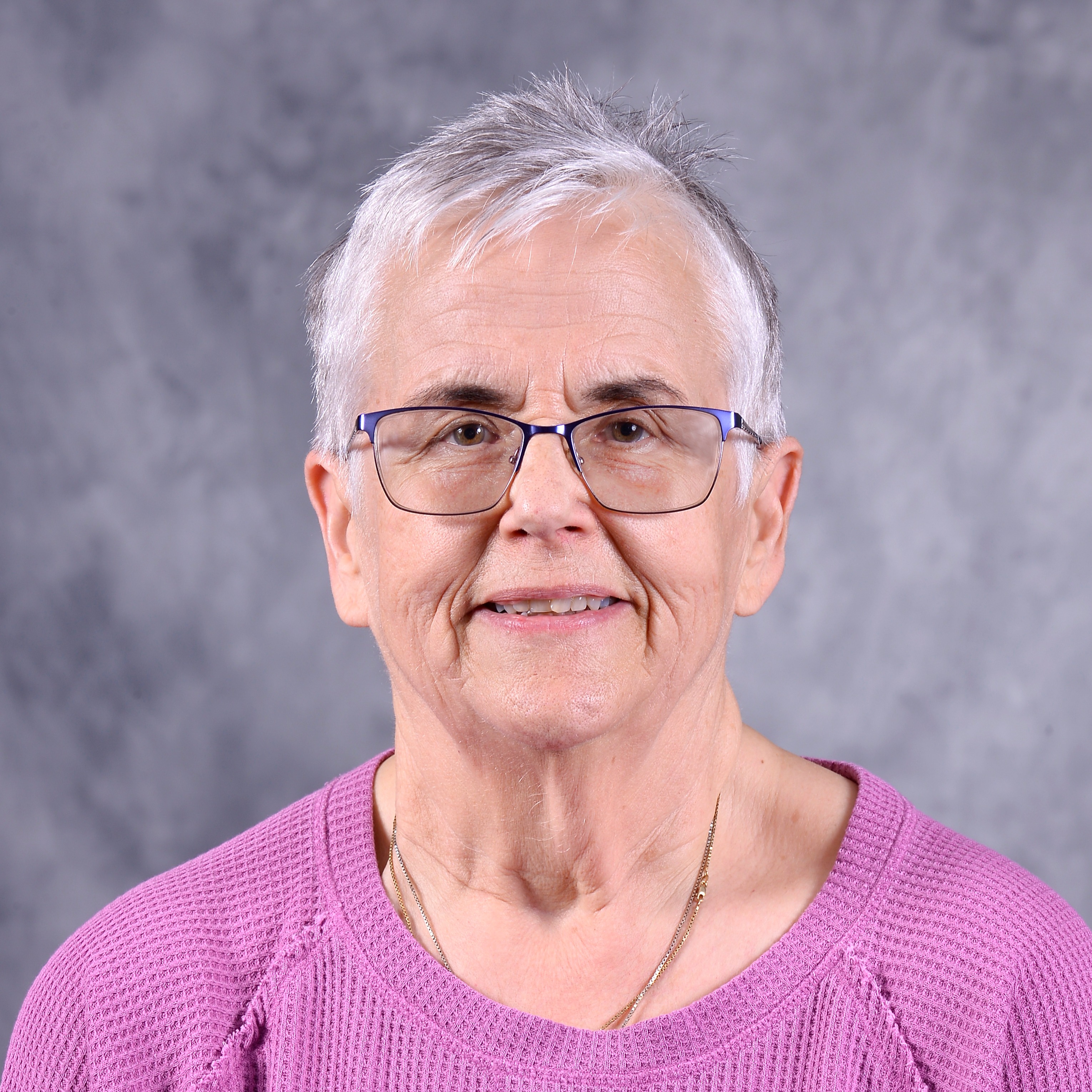 A smiling older woman with short gray hair and glasses, wearing a yellow blouse with floral embroidery, poses against a textured gray background.