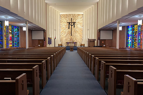 A view down the aisle of a modern church, featuring wooden pews, colorful stained glass windows on the sides, and a large cross at the altar against a sunburst backdrop.