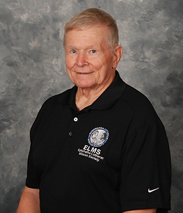 A smiling older man wearing a black polo shirt with a logo on the left side, standing against a textured gray background.