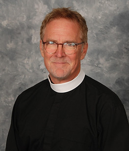 A man wearing glasses and a clerical collar, smiling against a textured gray background.