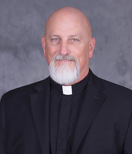 A man with a bald head and a white beard, wearing a black suit and a clerical collar, poses against a gray background.