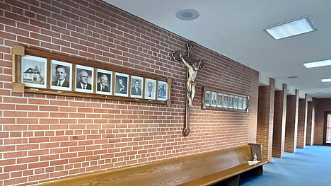 A brick wall featuring a series of framed portraits and a crucifix, with a wooden bench in the foreground and a well-lit corridor in the background.