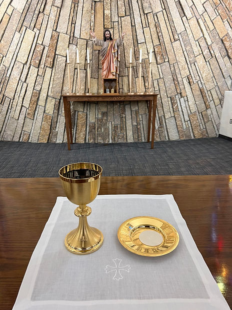 A golden chalice and a plate sit on a white cloth in the foreground, with a statue of Jesus in the background against a textured stone wall.