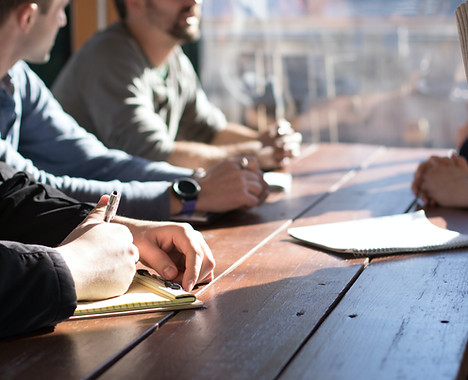 A group of people engaged in a discussion around a wooden table, with notebooks and pens visible, suggesting a collaborative meeting environment.