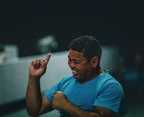 A man in a blue shirt is joyfully laughing and raising his hand in celebration, with a blurred background suggesting a lively atmosphere.