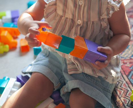 A young child is sitting on a patterned rug, holding colorful building blocks in their hands, with more blocks scattered around them.