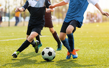 Two young soccer players in action, one in a black jersey and the other in a blue jersey, competing for the ball on a grassy field.