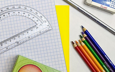 An assortment of school supplies including a protractor, graph paper, a yellow sheet of paper, colored pencils in various colors, and a silver pen arranged on a white surface.