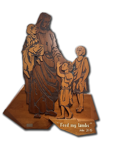 A wooden sculpture depicting a figure resembling Jesus with two children, one being held and the other reaching out, accompanied by the text 'Feed my lambs' and a biblical reference.