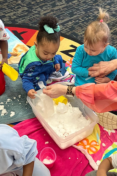 Two young children are engaged in a sensory play activity with a container filled with white material, surrounded by colorful blankets and toys.
