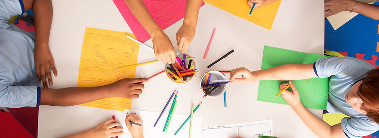 Aerial view of a table covered with colorful papers and art supplies, with several hands reaching for markers and crayons.