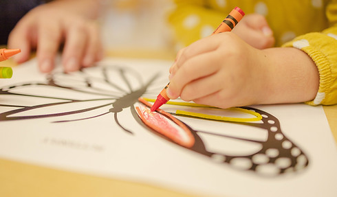 A child's hand is coloring a butterfly drawing with a red crayon, while other crayons are visible nearby.