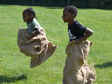 Two children are jumping in burlap sacks on a grassy field, enjoying a playful race.