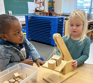 Two children are engaged in a hands-on activity with a wooden balance scale and various blocks in a classroom setting.