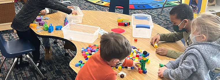 A group of children engaged in play at a table, using colorful building blocks and other materials in a classroom setting.
