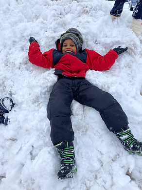 A child in a red jacket and black pants lies in a pile of snow, smiling and enjoying the winter weather.