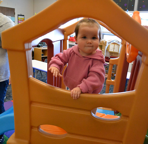 A young child in a pink hoodie is playfully peeking through an orange plastic play structure in a colorful indoor play area.
