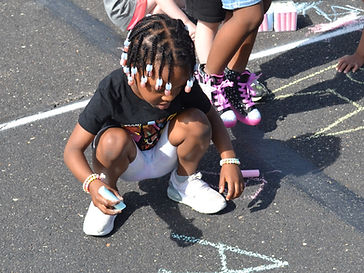 A young child with braided hair squats on a pavement, drawing with chalk while other children are visible in the background.