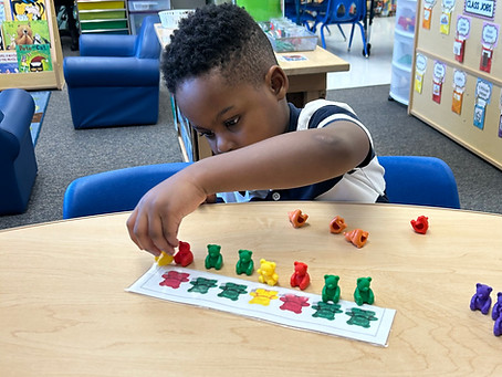 A young child is focused on arranging colorful bear-shaped counters in a specific order on a table in a classroom setting.