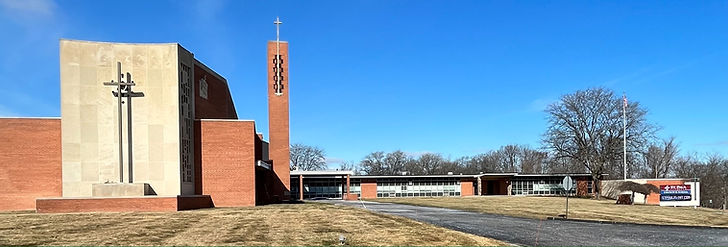 A view of a brick church building with a tall bell tower and a cross design on the facade, surrounded by a grassy area and clear blue sky.