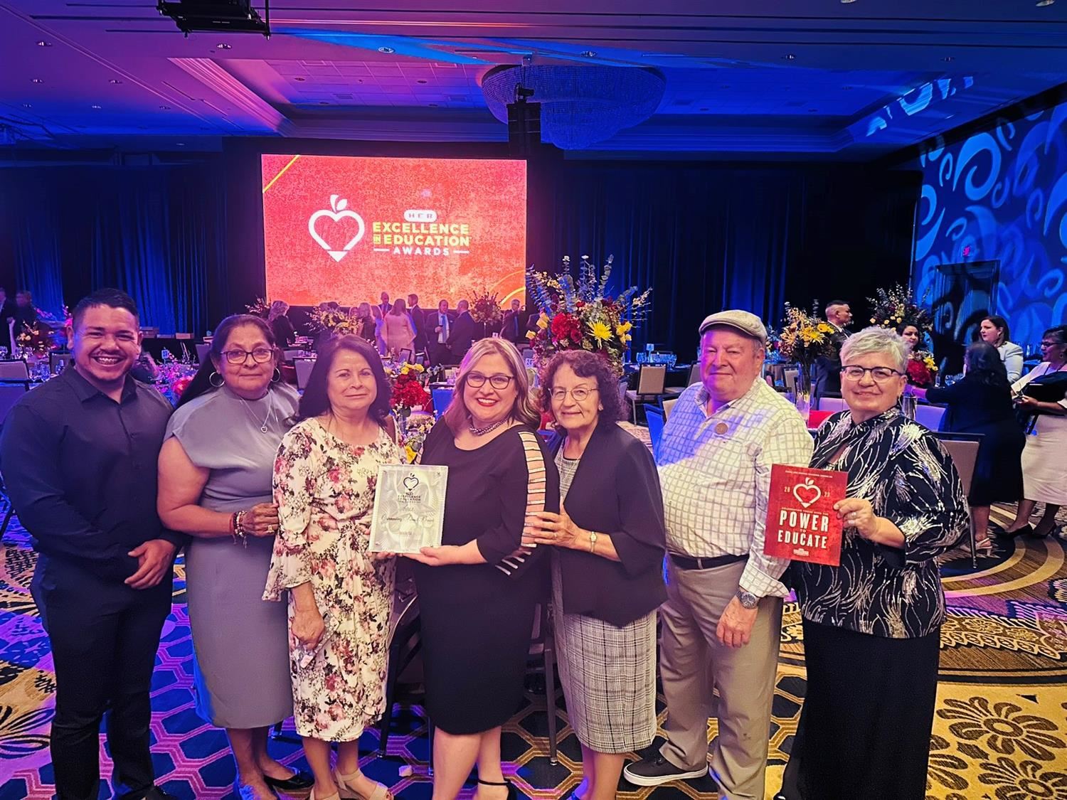A group of seven people stands together in a decorated venue, holding awards and smiling, with a large screen displaying a heart logo in the background.