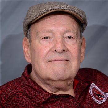 A senior man with a slight smile, wearing a flat cap and a maroon shirt, poses against a gray background.