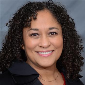 A woman with curly hair smiles warmly at the camera, wearing a dark jacket against a neutral background.