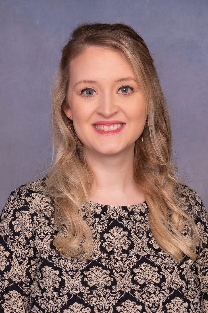 A woman with long, wavy blonde hair smiles at the camera, wearing a black and gold patterned dress against a blue background.