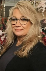 A woman with long, blonde hair and glasses poses in front of a festive background adorned with holiday decorations.