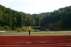 A view of a football field with goalposts in the foreground and a wooded hillside in the background under a clear sky.