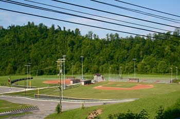A view of a baseball field surrounded by green hills, with two diamond-shaped fields, bleachers, and light poles under a clear blue sky.