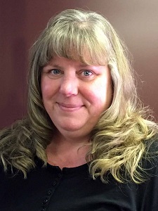 A woman with long, wavy blonde hair wearing a black top stands against a maroon background.