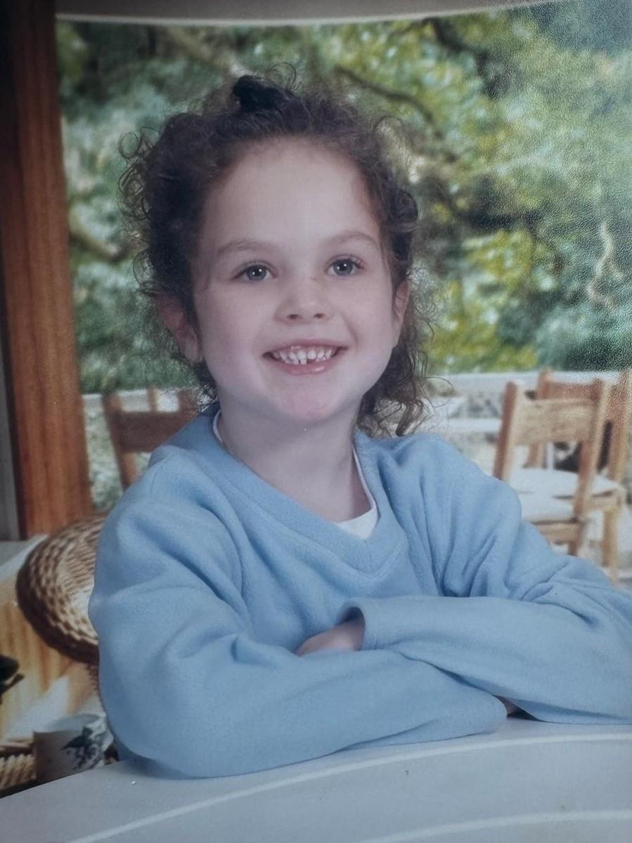 A young girl with curly hair smiles while sitting at a table, with a blurred outdoor background featuring trees and chairs.