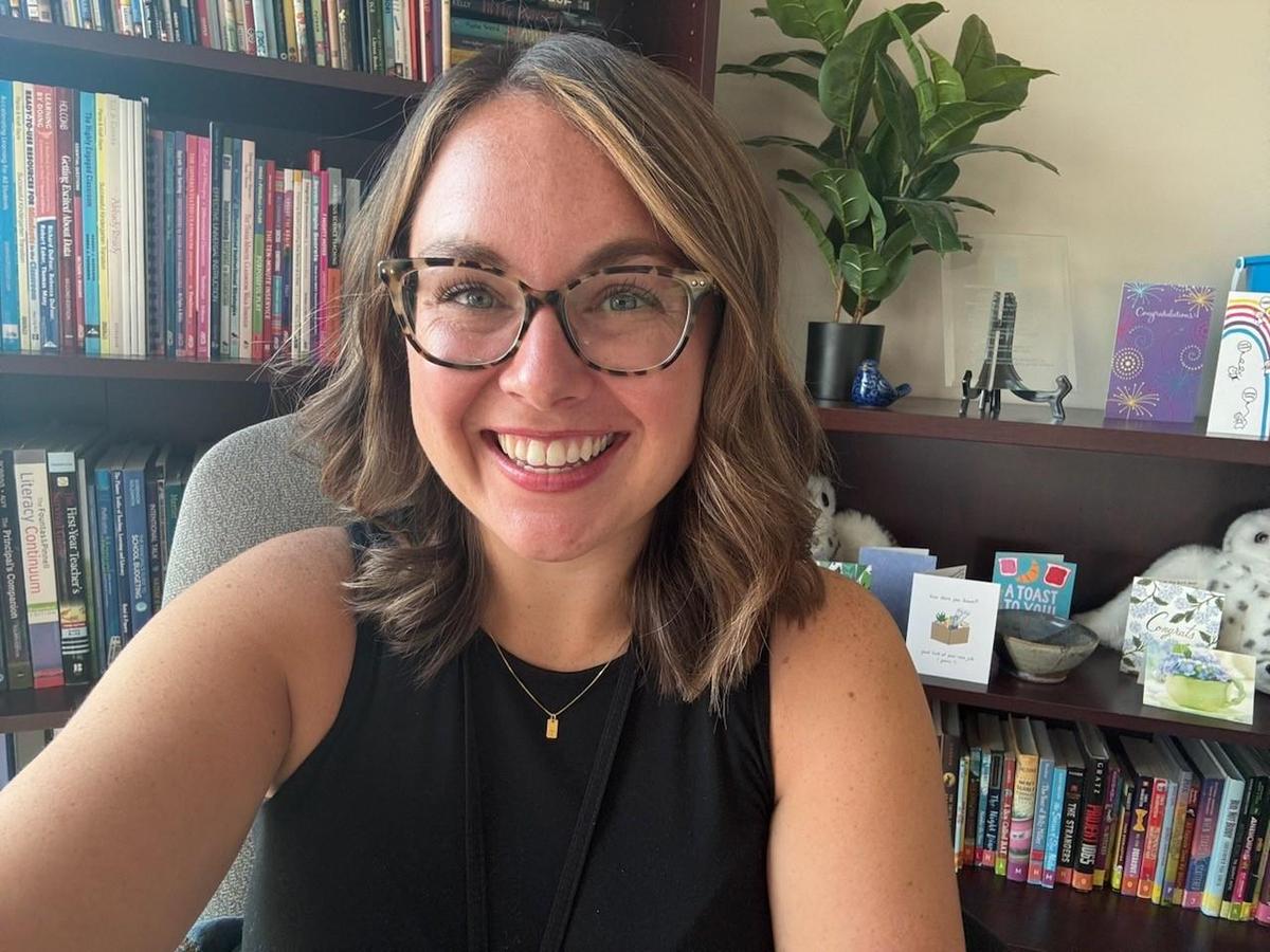 A smiling person with shoulder-length hair and glasses poses for a selfie in front of a bookshelf filled with books and decorative items, with a plant visible in the background.